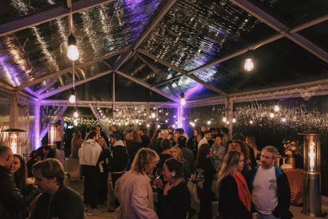 An evening party crowd mingling with drinks under a clear-roofed marquee strung with bulb lights.