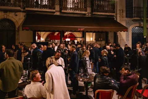A nighttime crowd holding wine glasses outside a Lisbon bistro with a red awning.