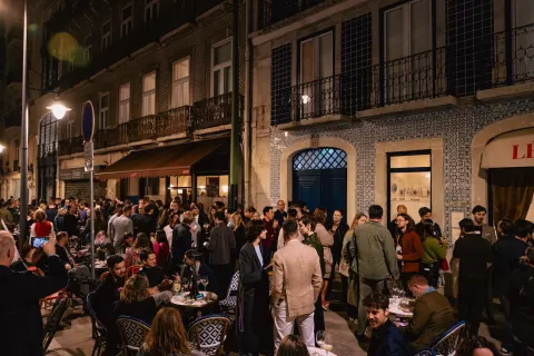 A nighttime crowd holding wine glasses outside a Lisbon bistro with a red awning.