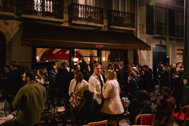 A nighttime crowd holding wine glasses outside a Lisbon bistro with a red awning.