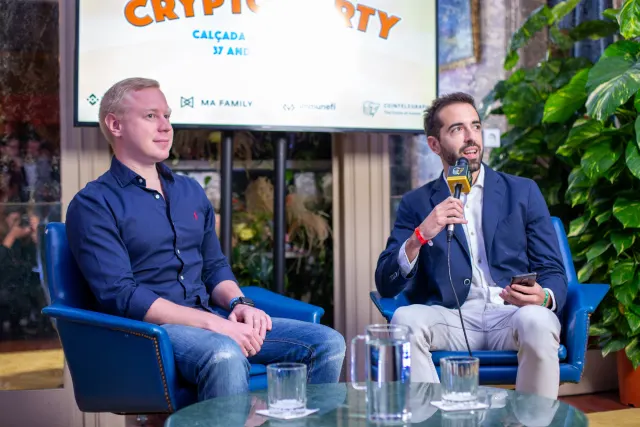Two men in a fireside chat on blue armchairs at Crypto Party, one holding a Cointelegraph microphone, sponsor logos on the screen behind.