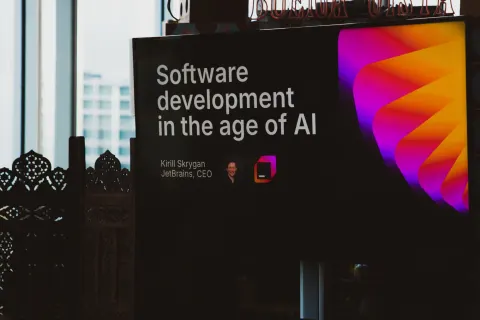A man presenting into a microphone to a small evening audience beside a window, a JetBrains slide on the screen at right.