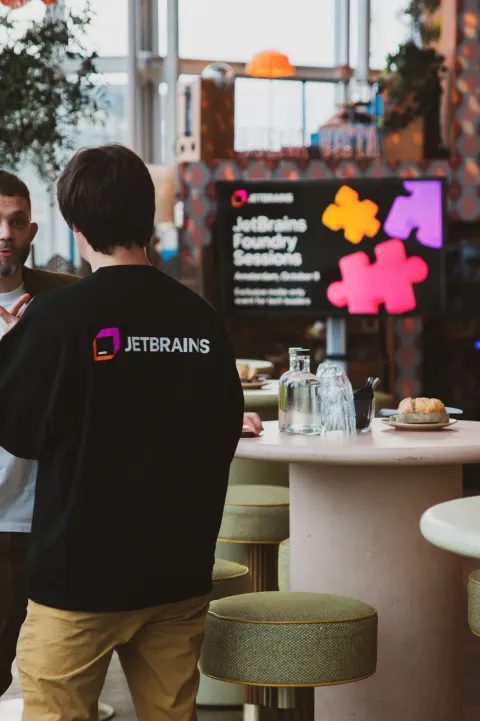 A man presenting into a microphone to a small evening audience beside a window, a JetBrains slide on the screen at right.
