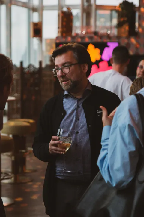 A man presenting into a microphone to a small evening audience beside a window, a JetBrains slide on the screen at right.