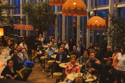 A man presenting into a microphone to a small evening audience beside a window, a JetBrains slide on the screen at right.