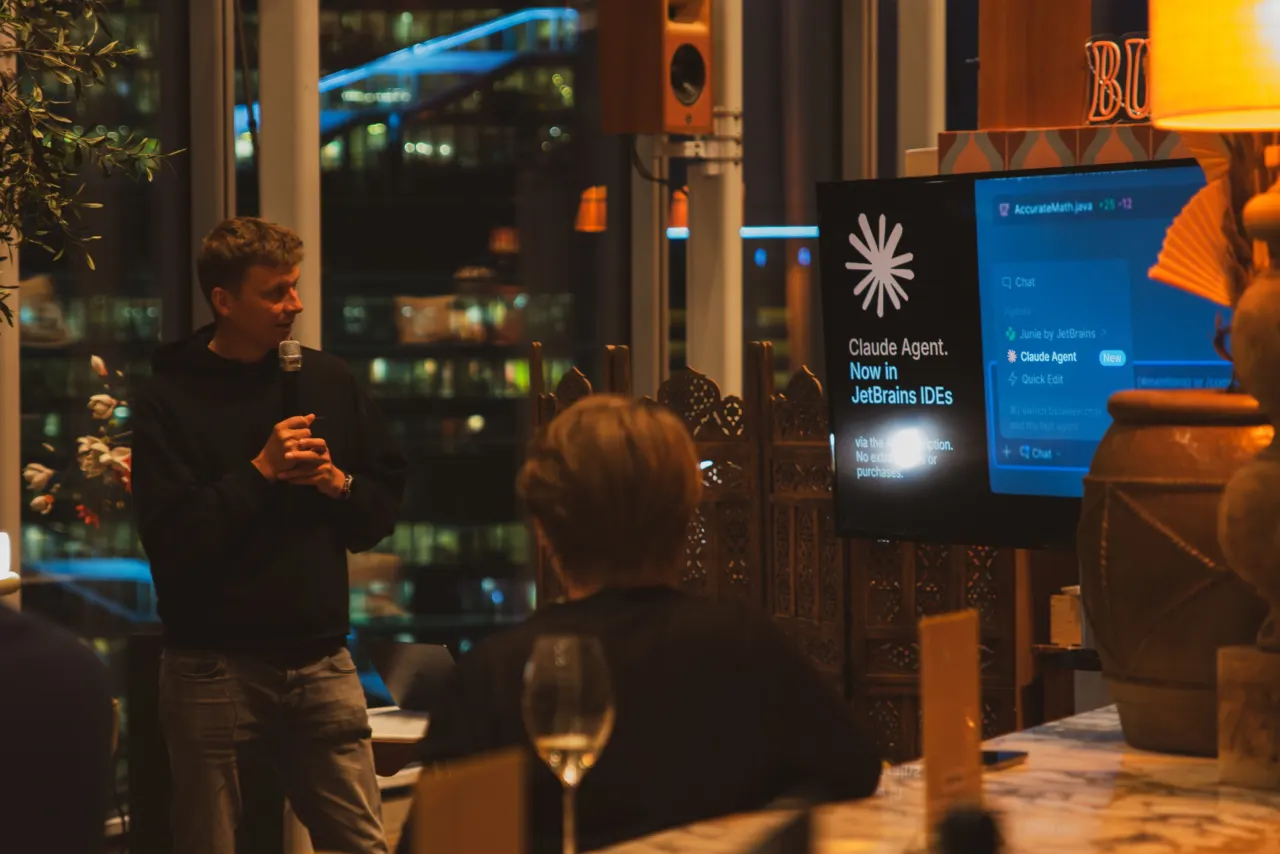 A man presenting into a microphone to a small evening audience beside a window, a JetBrains slide on the screen at right.