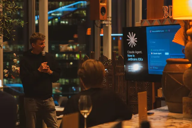A man presenting into a microphone to a small evening audience beside a window, a JetBrains slide on the screen at right.