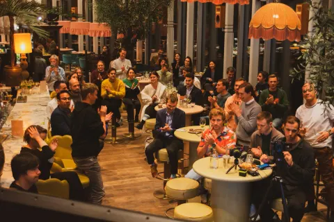 A man presenting into a microphone to a small evening audience beside a window, a JetBrains slide on the screen at right.