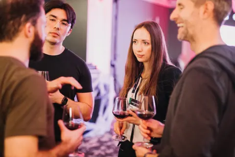 Four people talking and holding glasses of red wine at an event in a pink-lit room.