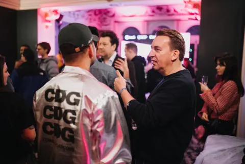 Four people talking and holding glasses of red wine at an event in a pink-lit room.