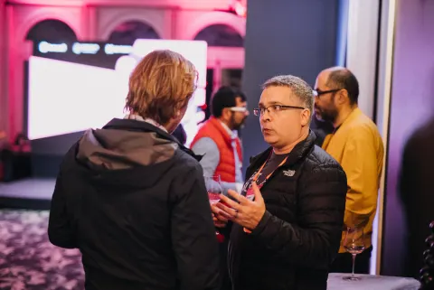 Four people talking and holding glasses of red wine at an event in a pink-lit room.