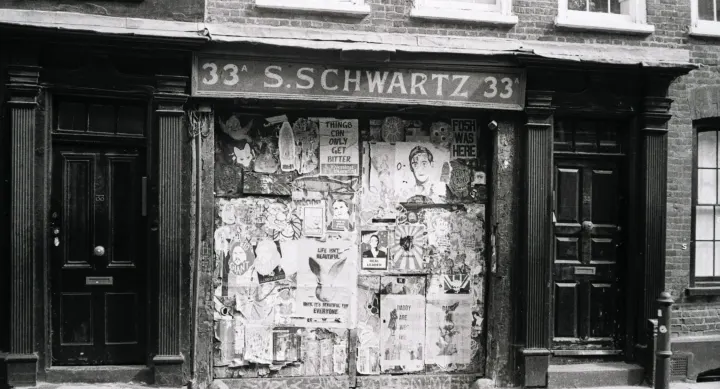 Black-and-white photo of an old boarded-up shopfront, 'S. Schwartz 33', covered with tattered posters.
