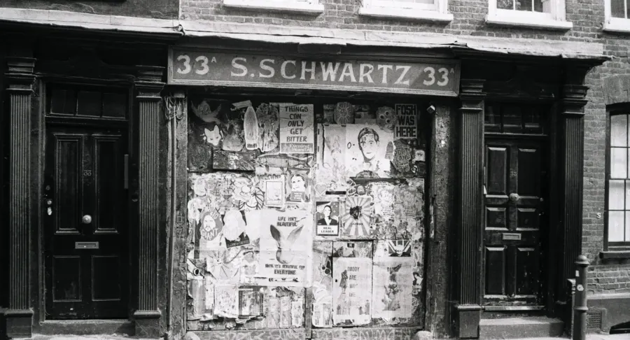 Black-and-white photo of an old boarded-up shopfront, 'S. Schwartz 33', covered with tattered posters.