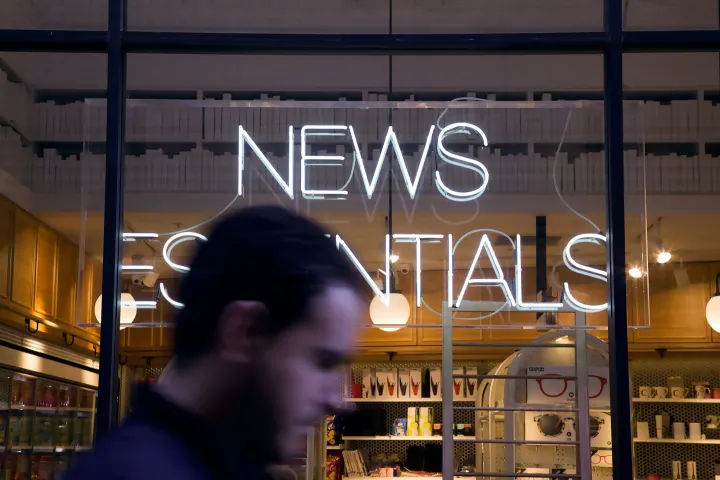 A man walking past a shop window with a glowing 'News Essentials' neon sign.