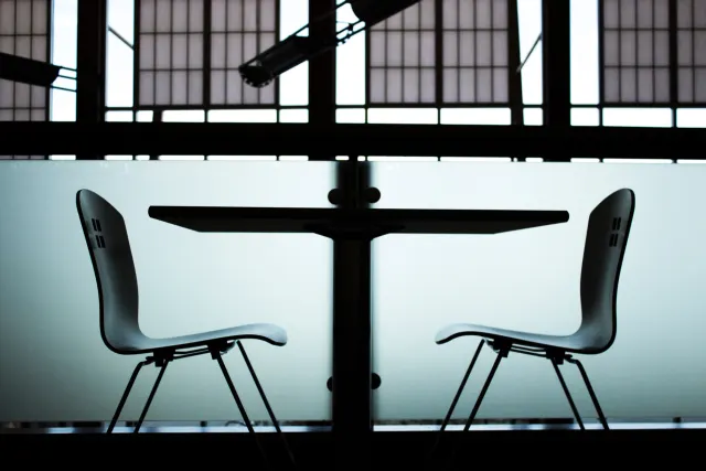 Two empty chairs facing each other across a small table in front of large windows, in silhouette.