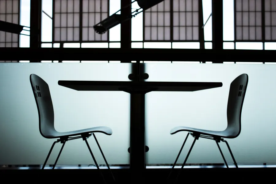 Two empty chairs facing each other across a small table in front of large windows, in silhouette.