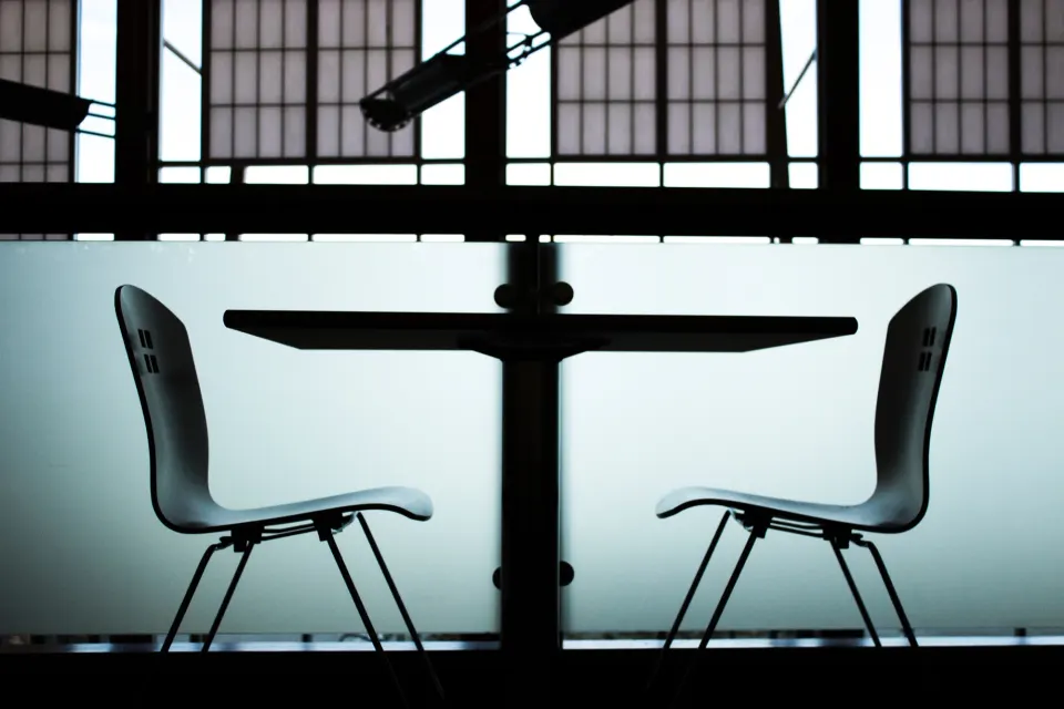 Two empty chairs facing each other across a small table in front of large windows, in silhouette.