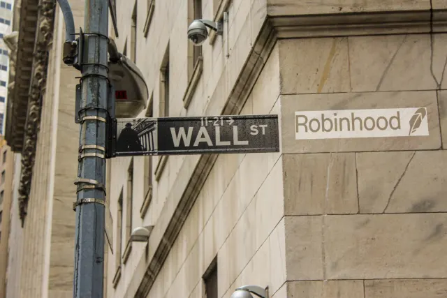 A 'Wall St' street sign beside a Robinhood logo sign on the corner of a stone building.
