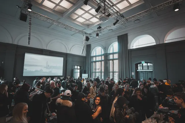 A large crowd of people gathered in a grand high-ceilinged hall with arched windows and a projection screen.