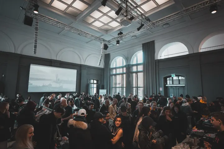 A large crowd of people gathered in a grand high-ceilinged hall with arched windows and a projection screen.