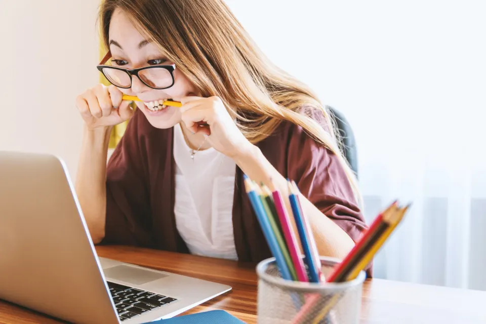 A woman in glasses biting a pencil while staring at a laptop, colored pencils in the foreground.