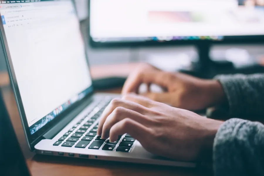 Close-up of hands typing on a laptop at a desk with a second monitor behind.